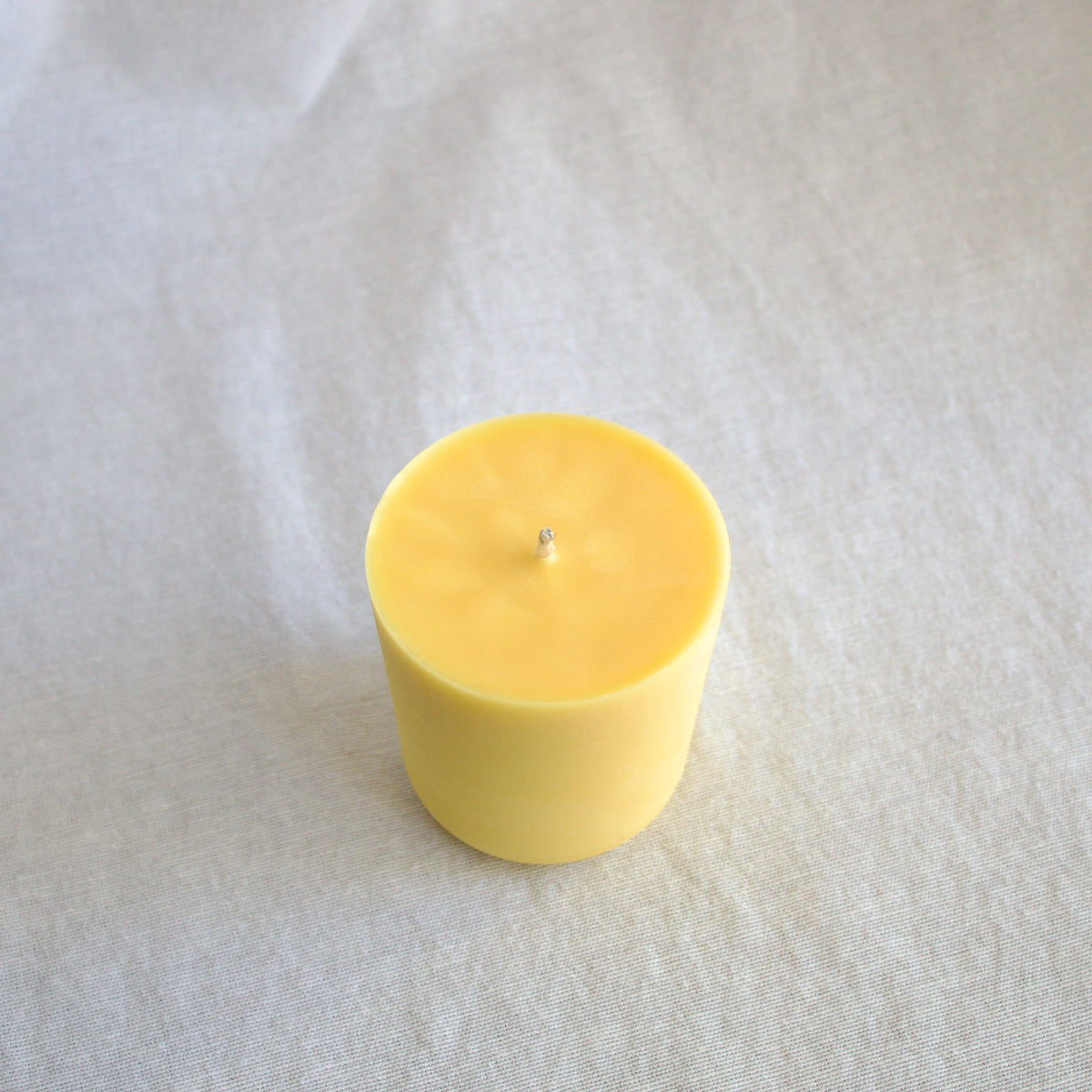 A refillable beeswax candle with a small cotton wick, centered on a textured off-white fabric background.