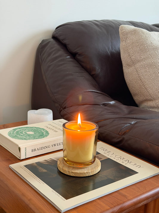 A lit candle in a clear glass jar sits on a wooden coaster on a wooden table, styled in a cozy living room setting next to a brown leather sofa and a magazine.