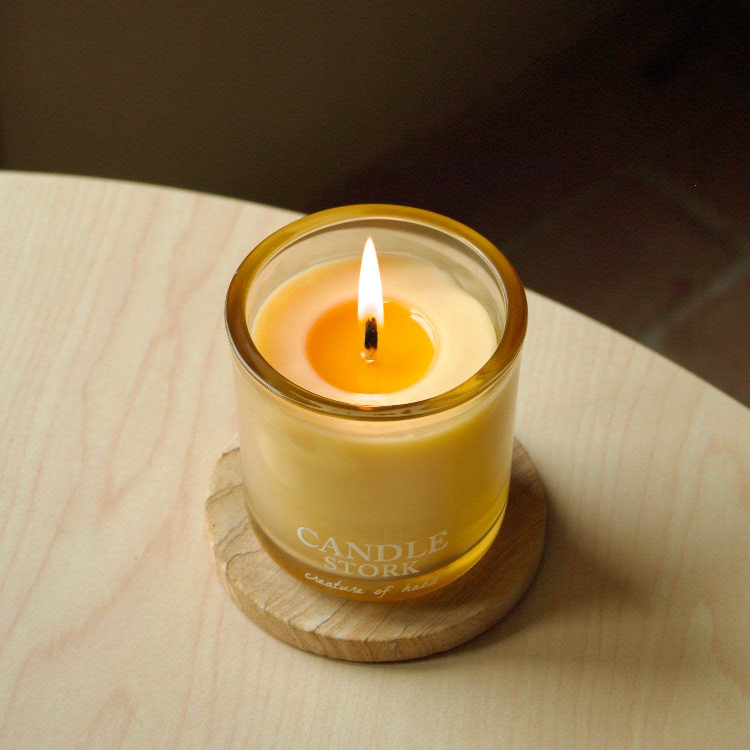 A lit candle in a clear glass jar with a gold-toned rim and pale yellow wax, sitting on a round wooden coaster on a light wood tabletop. The candle label reads CANDLE STORK.
