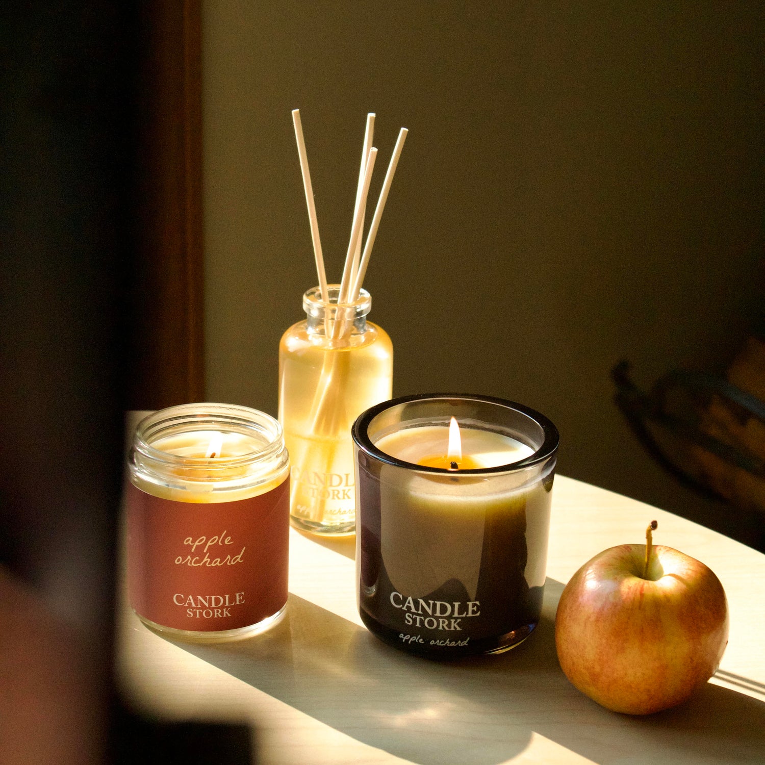 Two lit candles and a reed diffuser set on a table in warm sunlight, with a red apple beside them. Visible labels include CANDLE STORE on the black glass candle jar and apple orchid on the amber glass candle jar.