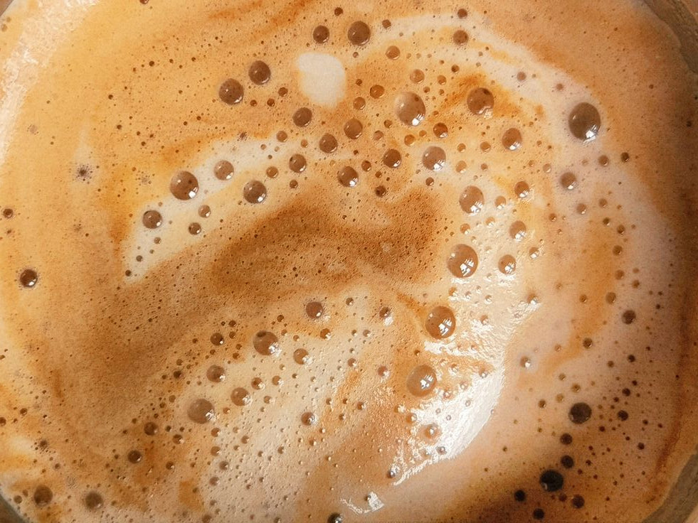 A close-up top-down view of a frothy latte with swirling layers of creamy beige foam and golden bubbles.
